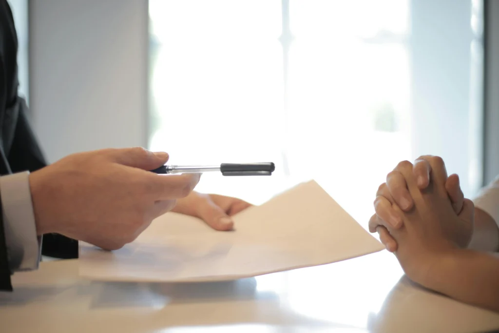 A banker holds a contract in one hand and a pen in the other and hands both to the customer for her to sign